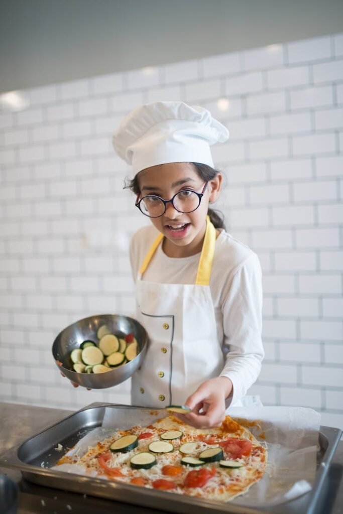 Child chef in apron and toque assembling pizza with fresh zucchini in a kitchen.