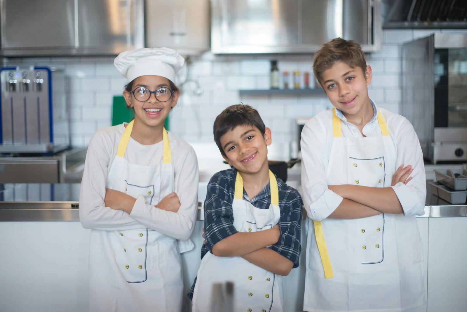 Three smiling children in aprons pose in a professional kitchen.