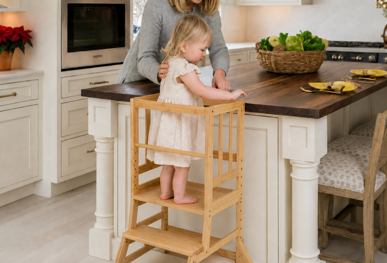 Toddler standing safely in a bamboo kitchen learning tower while an adult supervises at a kitchen island.